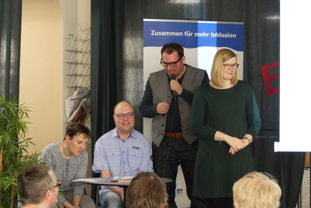 Von links: Am Tisch sitzen Maria Popp und Oliver Kuhn von der Prüfergruppe Alles klar. Volker Glombitza leitet die Prüfergruppe und erzählt gerade von der gemeinsamen Arbeit. Rechts steht die Gebärdensprachdolmetcherin Kathleen Entrich.